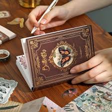 A person's hands holding a vintage-style illustrated notebook or card deck on a wooden table, surrounded by scattered cards and stationery.
