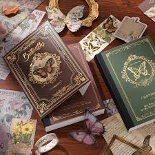 A styled flat-lay of vintage-style decorative books and stationery on a wooden surface. The main products appear to be ornate books with butterfly illustrations on the covers, surrounded by matching cards, pins, butterflies, and paper props.