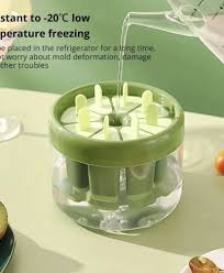 A clear silicone ice cream stick mold with green sticks, placed in a green box, with a green lid, and a glass of popsicles in the background.