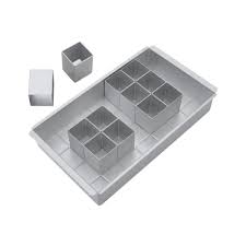 A silver-colored adjustable baking pan with grid grooves on the bottom, along with four small white square molds for shaping cakes.